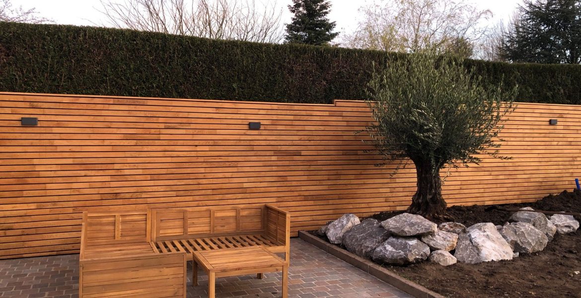 Une réalisation de la société Langlet dans un jardin de particulier dans l'Oise. Une clôture composée de lames en bois horizontales et un massif de plantes contenant un bel olivier entouré de pierres. Derrière la clôture se trouve une haire venant d'être taillée