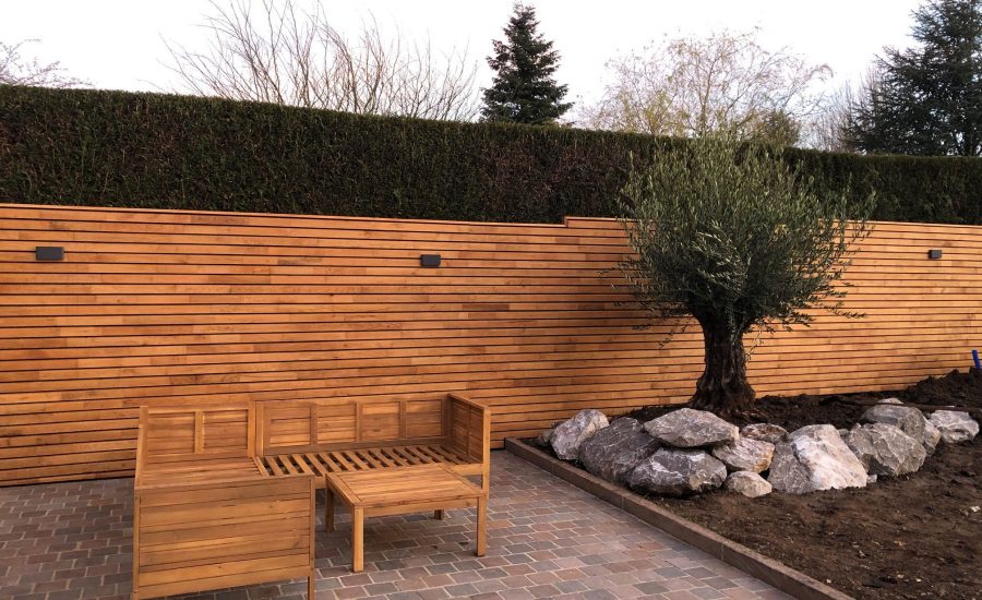Une réalisation de la société Langlet dans un jardin de particulier dans l'Oise. Une clôture composée de lames en bois horizontales et un massif de plantes contenant un bel olivier entouré de pierres. Derrière la clôture se trouve une haire venant d'être taillée