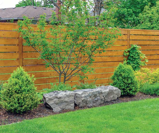 une clôture de panneau rigides en bois et un massif d'arbustes verdoyants et trois grosses pierre dans le jardin d'un particulier dans l'Oise. On voit aussi du beau gazon